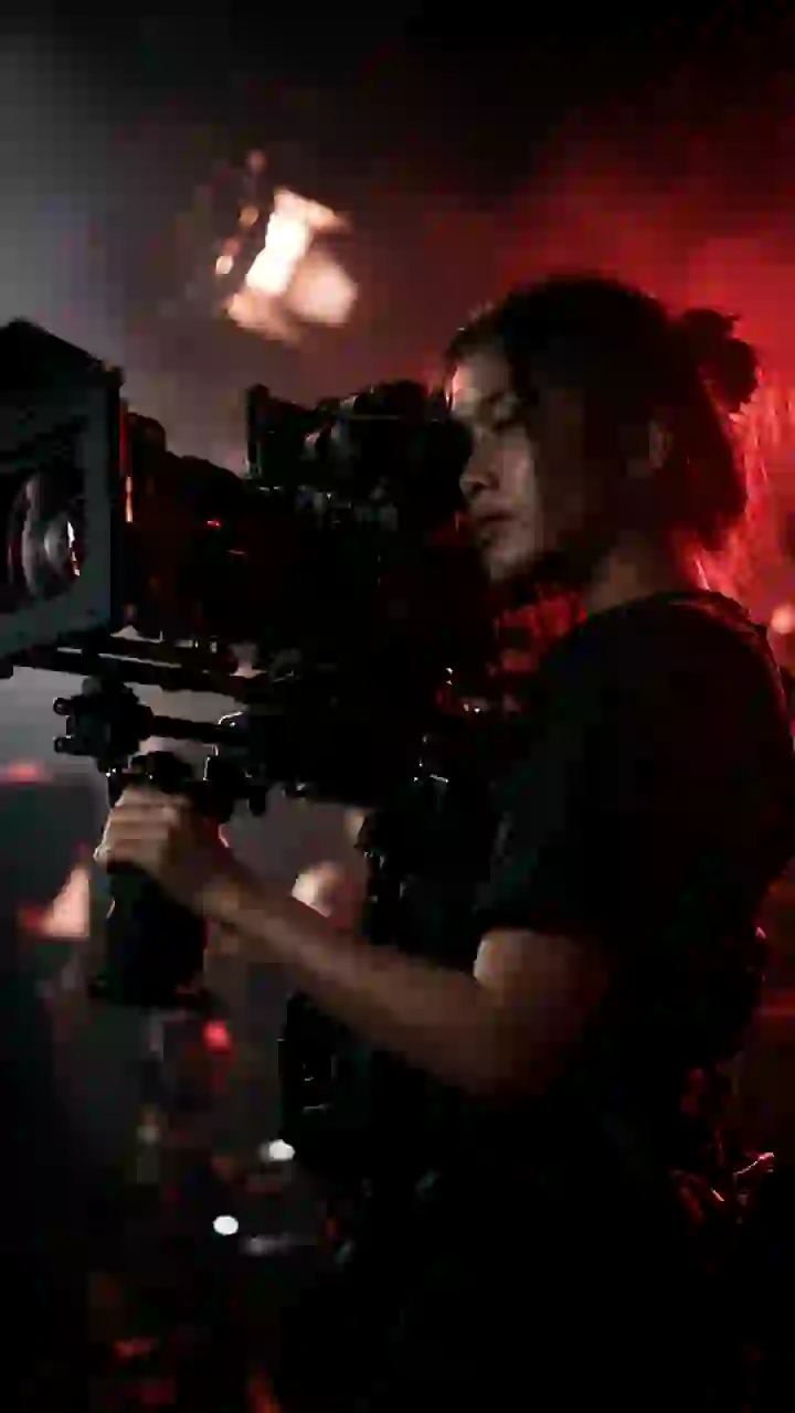 A camera operator with a shoulder-mounted cinema camera in red rim light.