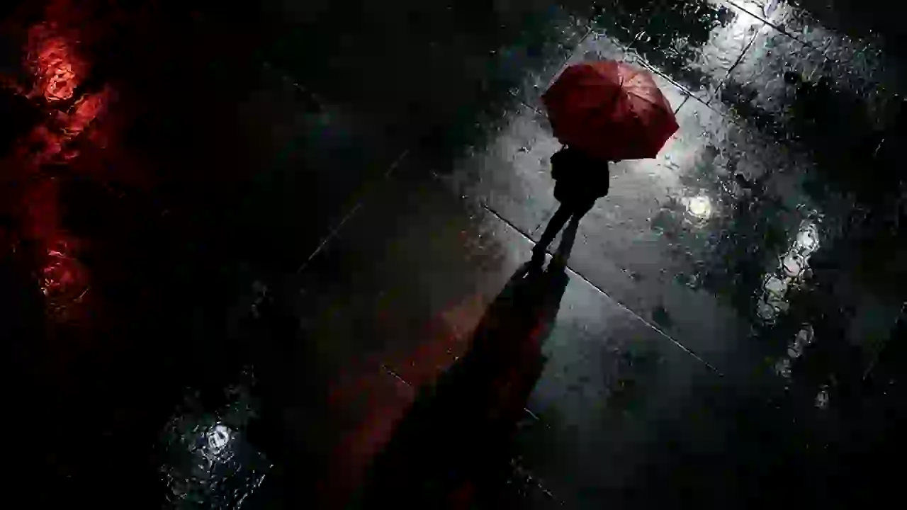 Overhead shot: a lone person with a red umbrella crosses a wet black square at night.