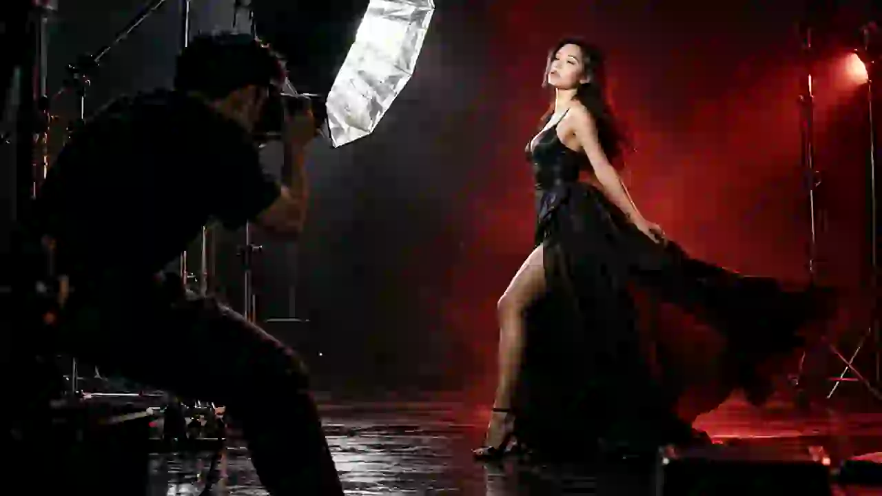 A photographer fires a flash at a model in a long black dress on a dark set.