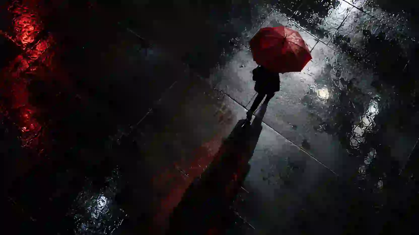 Overhead shot: a lone person with a red umbrella crosses a wet black square at night.