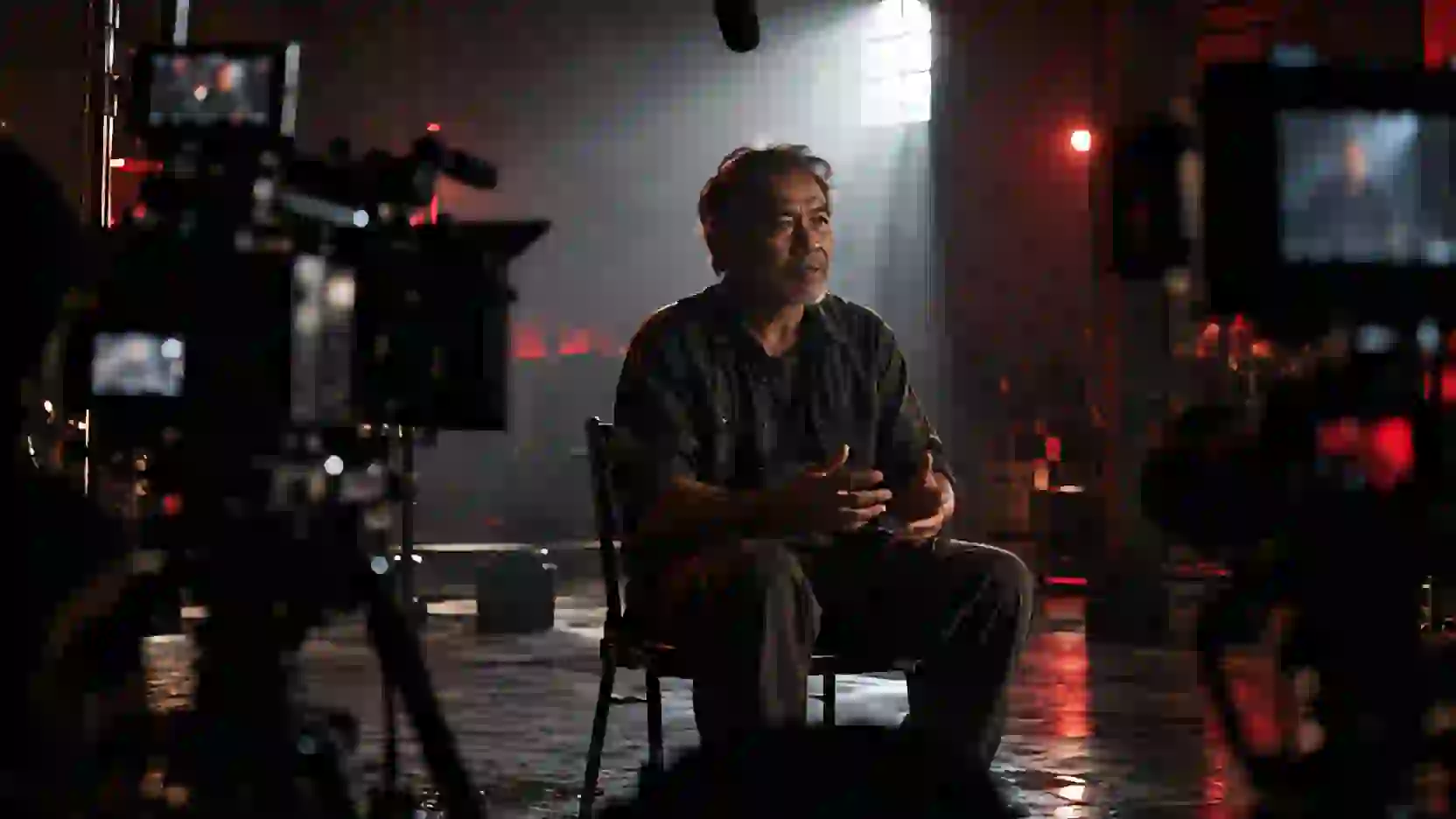 Documentary interview: an older man speaks in a dark hall, backlit, two cameras in the foreground.