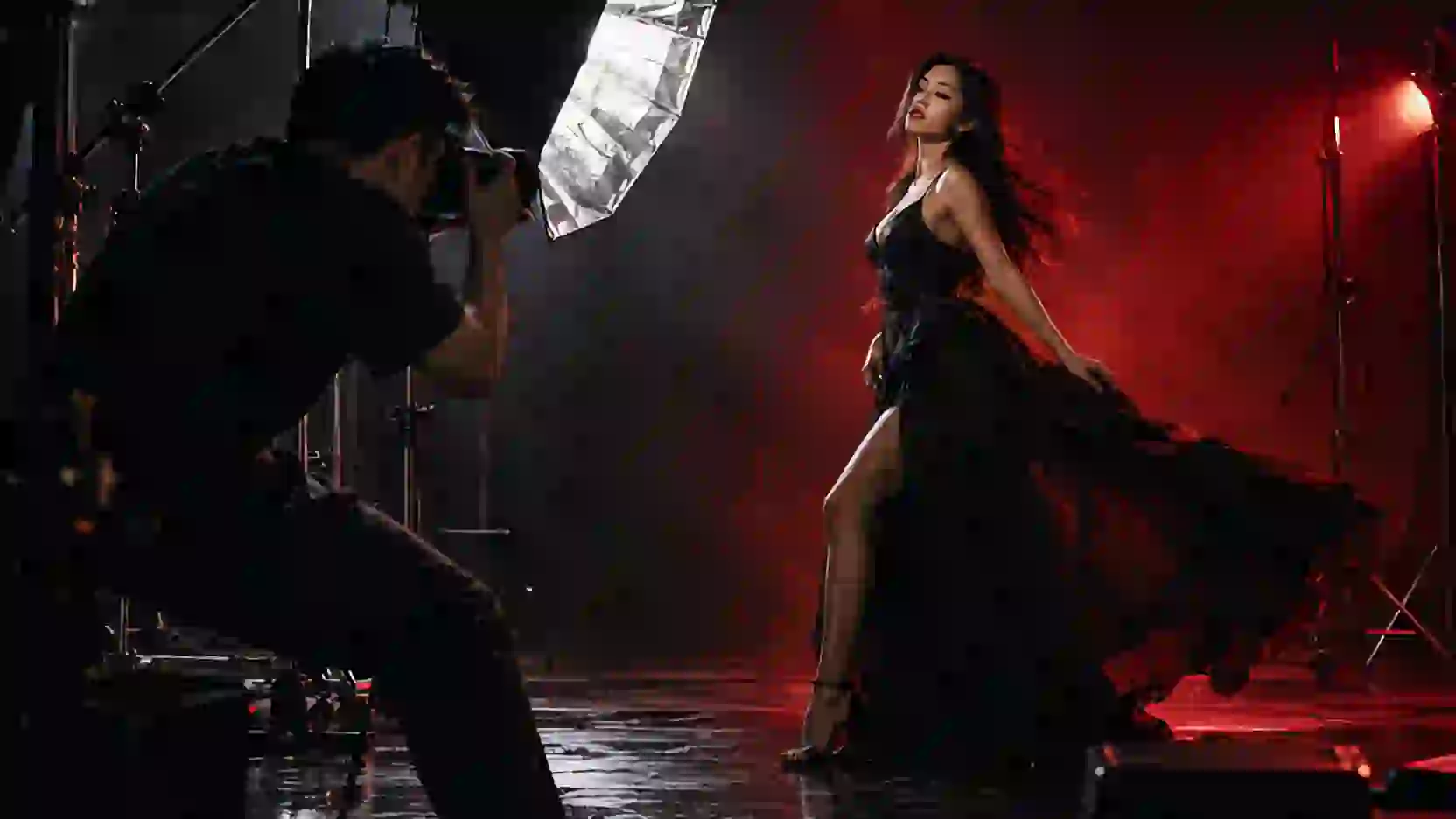 A photographer fires a flash at a model in a long black dress on a dark set.